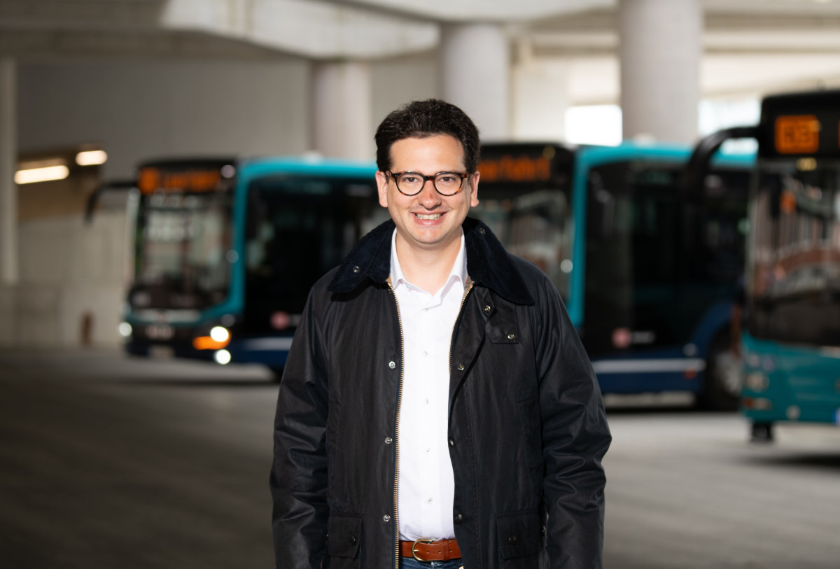 Bastian Mahmoodi steht in einem Depot mit Linienbussen. Er blickt zentral in die Kamera.