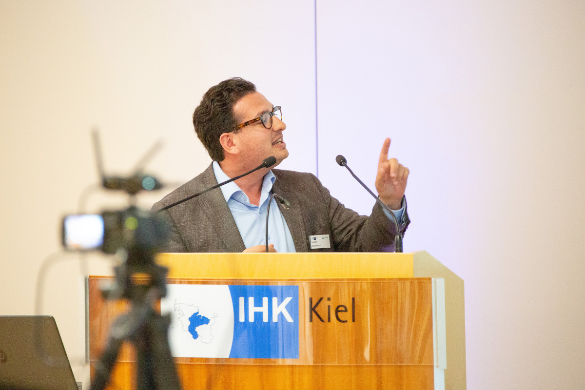 Bastian Mahmoodi spricht bei einer Veranstaltung der IHK Kiel am Rednerpult und gestikuliert während seines Vortrags.