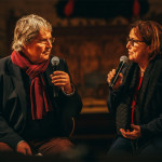 Zwei Personen führen ein Bühnengespräch mit Mikrofonen vor Publikum.