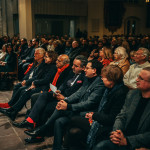 Bastian Mahmoodi sitzt mit weiteren Personen in einem Veranstaltungsraum.