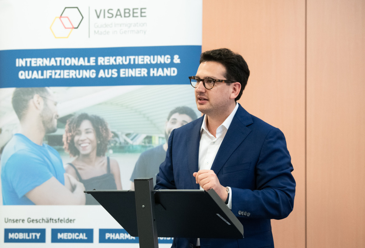 Bastian Mahmoodi spricht an einem Rednerpult vor einem VISABEE-Banner über internationale Fachkräfterekrutierung.