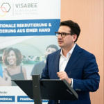 Bastian Mahmoodi spricht an einem Rednerpult vor einem VISABEE-Banner über internationale Fachkräfterekrutierung.